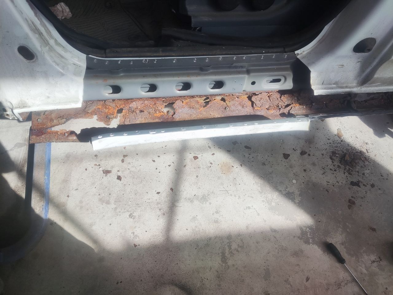 Zniszczony, skorodowany próg samochodu przed naprawą. Widoczne ślady rdzy i uszkodzeń blacharskich. Fragment karoserii w kolorze białym.