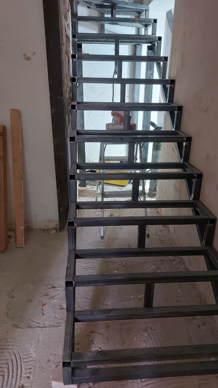 Metalowa konstrukcja schodów w surowym stanie, spawana z profili kwadratowych, widoczna na tle ściany w trakcie budowy.
