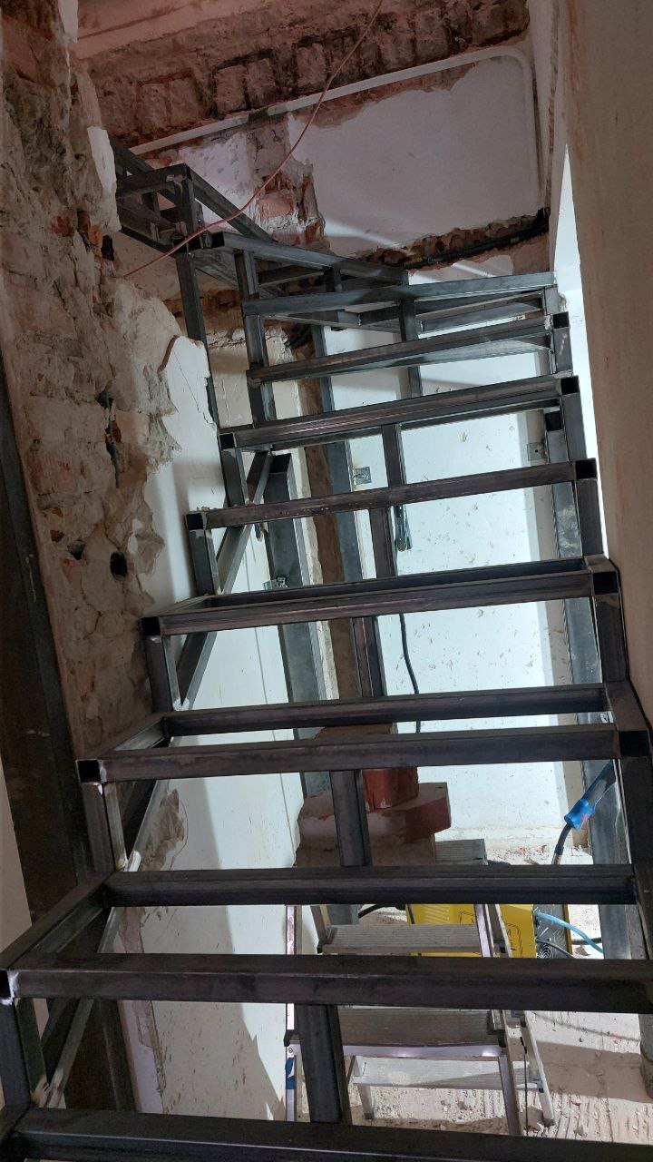 Metalowa konstrukcja schodów w surowym stanie, spawana, z widocznym sprzętem spawalniczym, w trakcie montażu w budynku z odsłoniętą cegłą i fragmentami tynku.