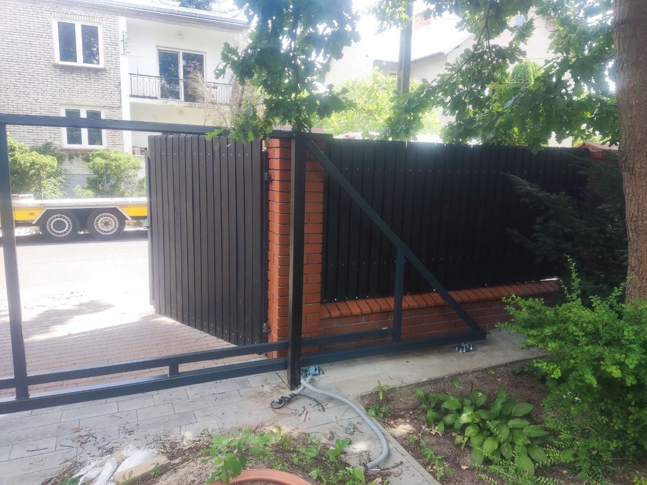 Nowoczesna brama wjazdowa przesuwna z ciemnego drewna i ceglanymi słupkami, widoczna z boku na tle domu i drzew. Elementy metalowe w kolorze grafitowym.