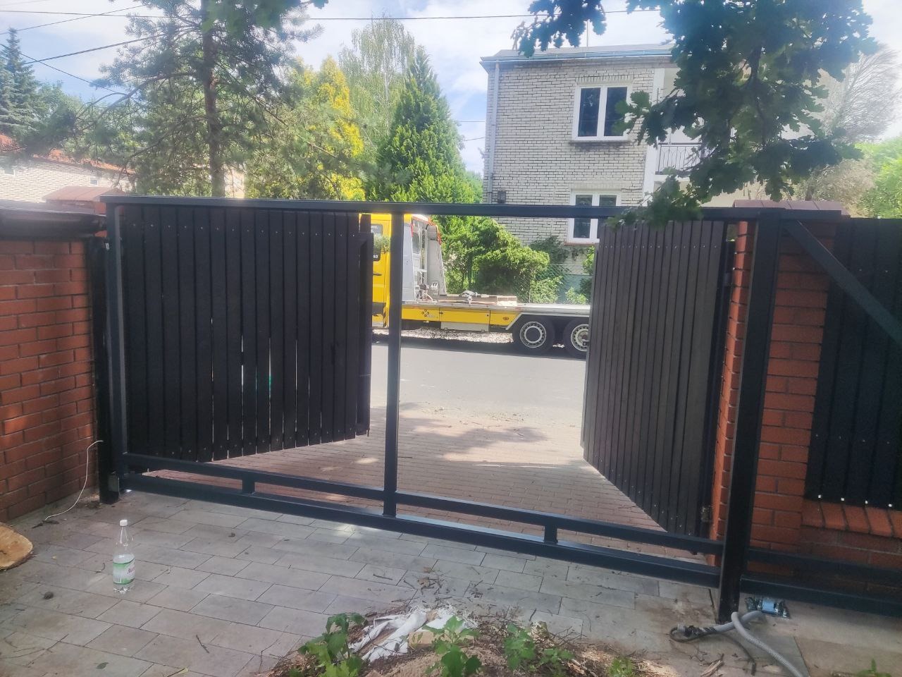 Otwarta, nowoczesna brama wjazdowa z ciemnego drewna i metalową ramą, na tle domu z cegły i zieleni. Widoczna laweta w tle.