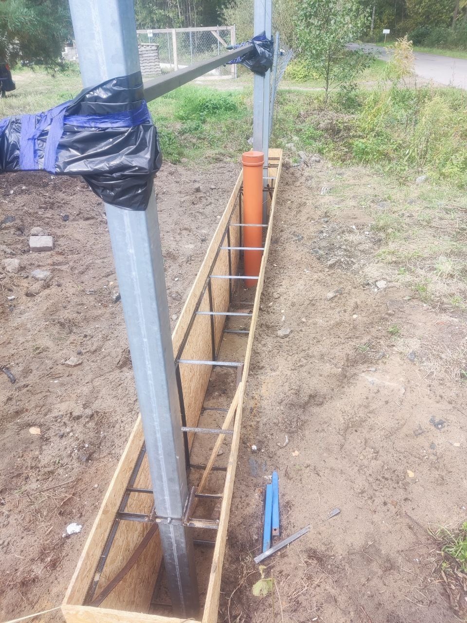 Metalowe słupy w drewnianej szalunku, przygotowane do zalania betonem. Widoczna pomarańczowa rura. Prace ziemne w toku, teren trawiasty w tle.