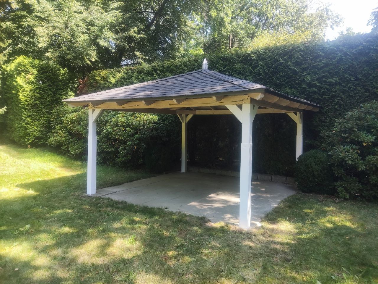 Biała, drewniana altana ogrodowa z szarym dachem, otoczona zielenią trawnika i żywopłotu. Widok na konstrukcję w słoneczny dzień.