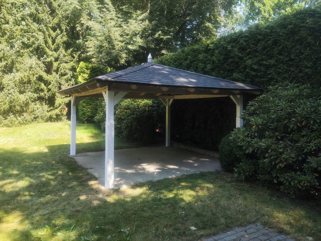 Zadaszona altana ogrodowa z białymi słupami i ciemnym dachem, otoczona zielenią. Widok z zewnątrz, w słoneczny dzień. Konstrukcja wkomponowana w krajobraz.