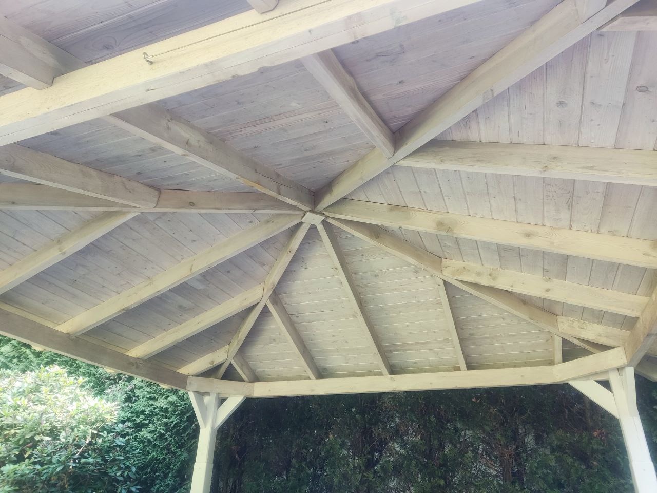 Drewniana konstrukcja zadaszenia altany ogrodowej, widok od dołu. Jasne drewno, regularny układ belek i desek. W tle zieleń drzew i krzewów.