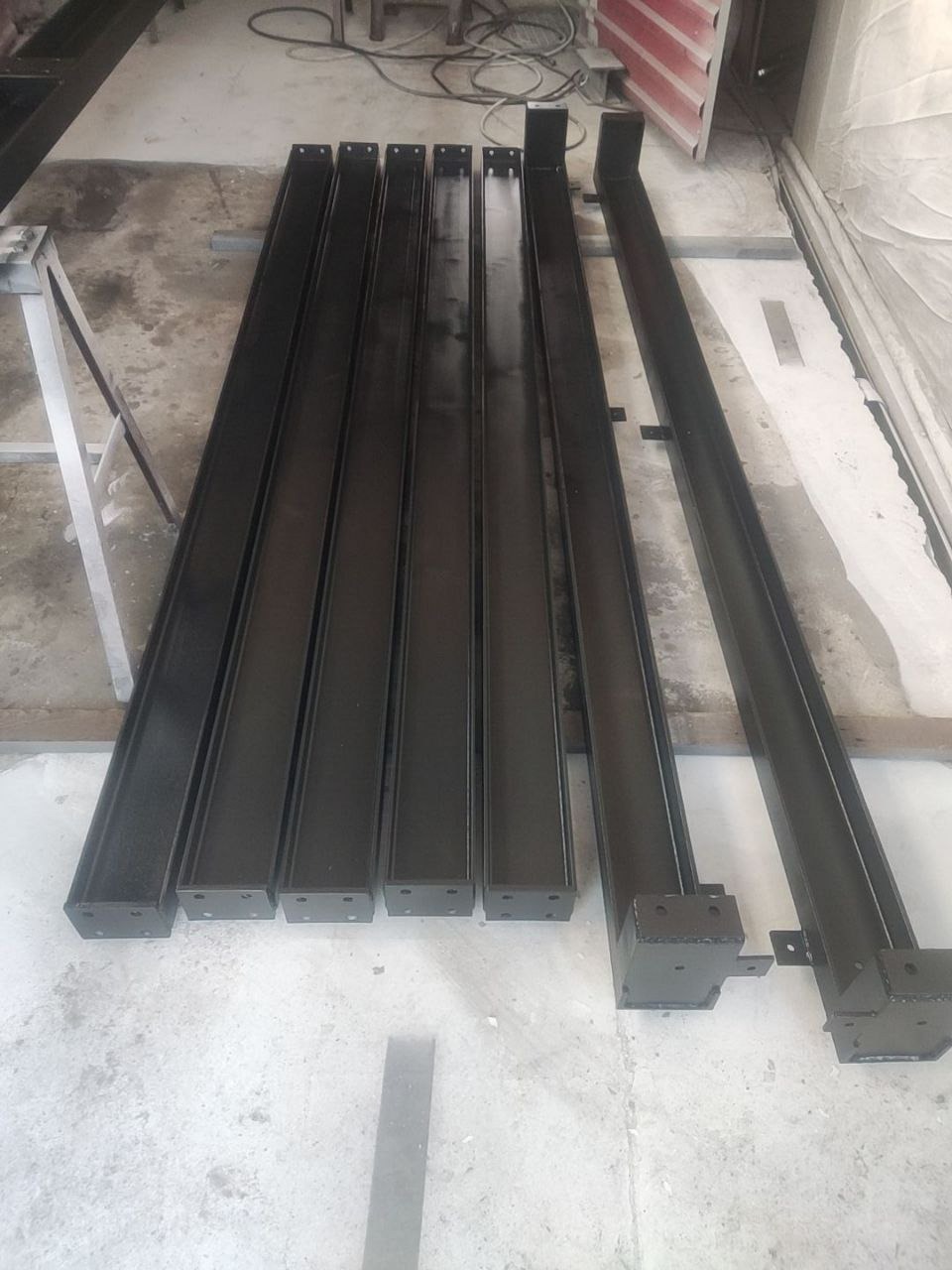 Czarne, metalowe profile o przekroju prostokątnym, ułożone równolegle na szarym, betonowym podłożu. Widoczne otwory montażowe i spawane elementy.