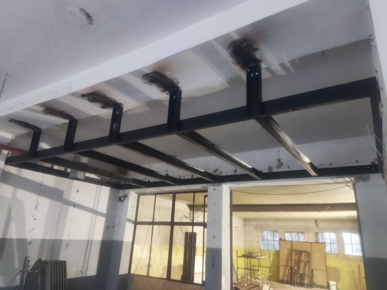 Metalowa konstrukcja podwieszana do sufitu, spawana, pomalowana na czarno. Widoczne spawy i mocowania do sufitu. W tle okna i elementy wyposażenia warsztatu.