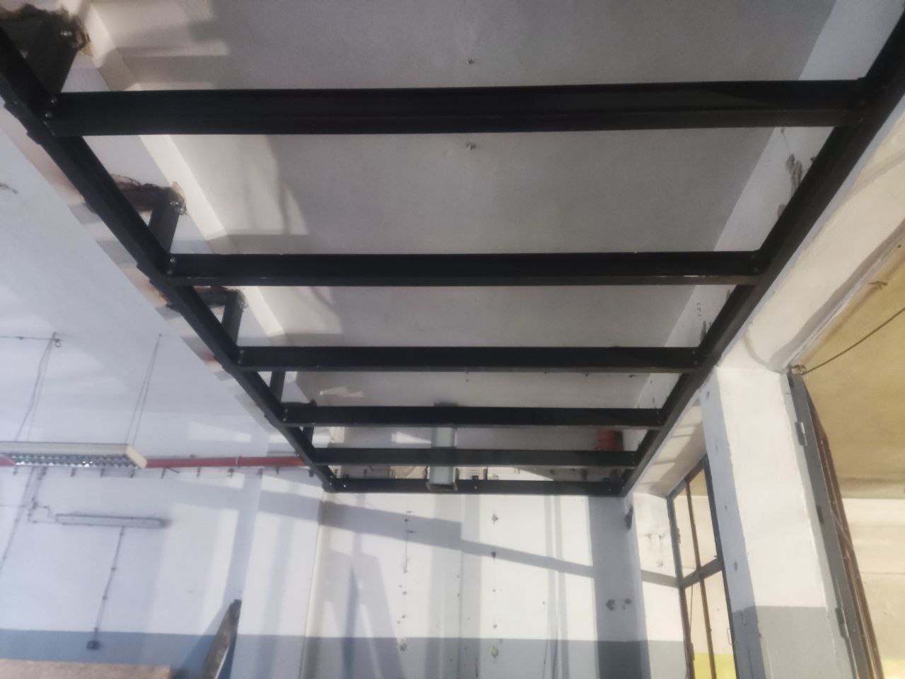 Czarna, metalowa konstrukcja sufitowa z profili, widoczna z dołu, w jasnym pomieszczeniu. Elementy spawane, surowy wygląd, widoczne ślady montażu.