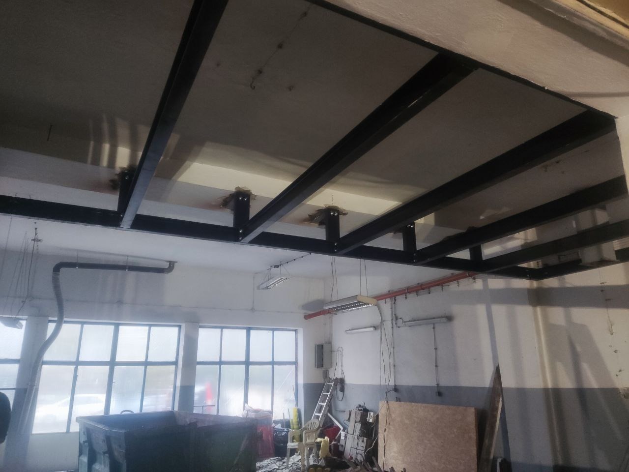Metalowa konstrukcja podwieszana pod sufitem w hali, widoczne spawane elementy i mocowania. W tle okna i wyposażenie warsztatu.