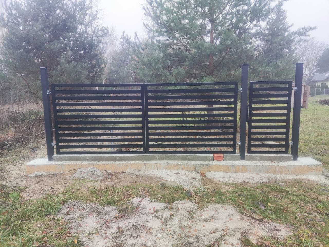 Nowoczesna, czarna brama wjazdowa z poziomymi szczeblami, osadzona na betonowej podstawie. W tle drzewa i trawa, widoczne detale montażu.