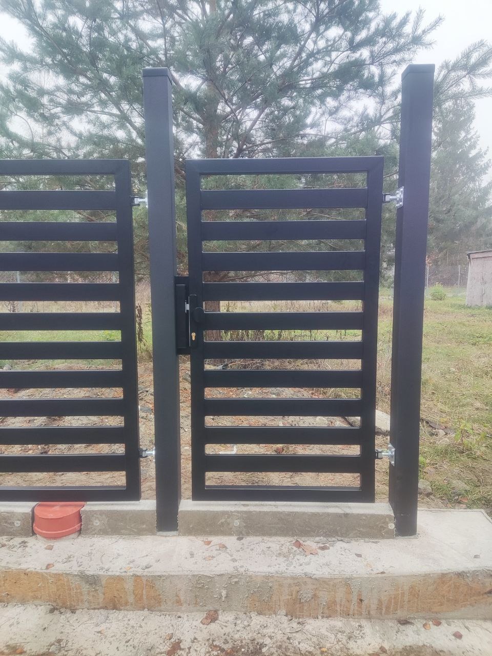 Nowoczesna, czarna brama wjazdowa z poziomymi szczeblami, zamontowana na betonowej podstawie. Widoczne słupki i zawiasy. Tło stanowi zieleń ogrodu i drzewa.