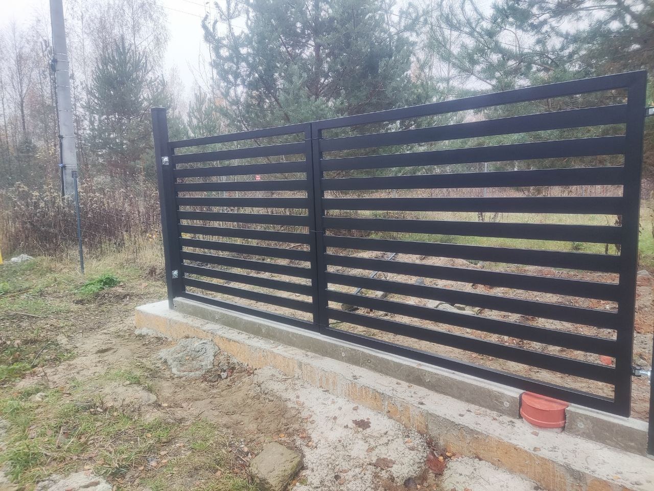 Nowoczesna brama wjazdowa z poziomych profili, pomalowana na czarno, zamontowana w Pruszkowie. Widoczne betonowe fundamenty i otoczenie posesji.