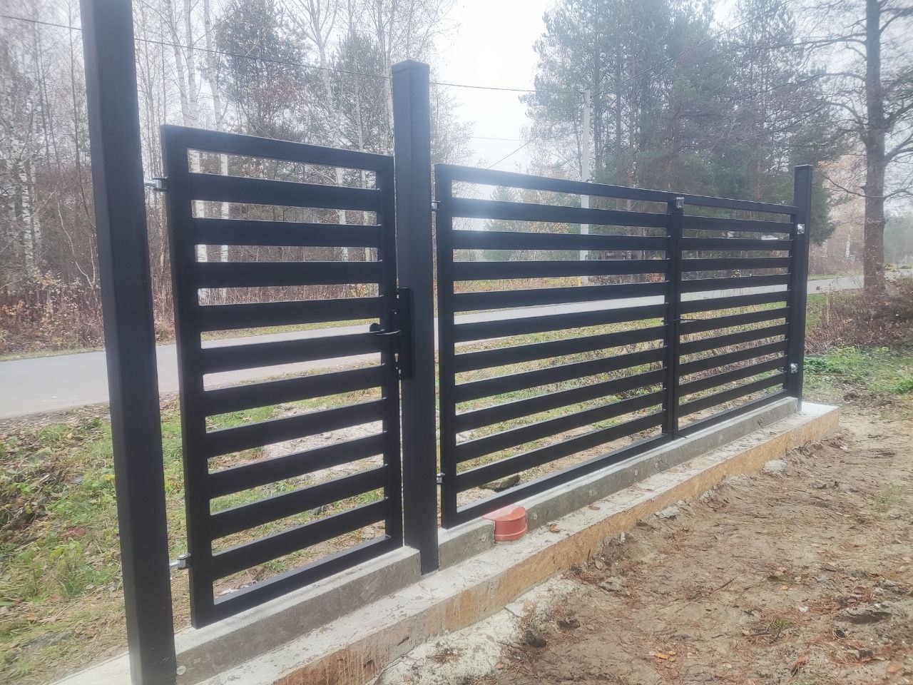 Nowoczesna, czarna brama wjazdowa z poziomymi szczeblami, osadzona na betonowej podstawie. Tło stanowią drzewa i droga.