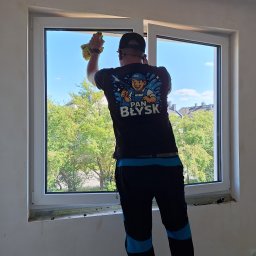 Adam Maksylewicz - Pracownik w Szczecinie myje okno, widok z tyłu. Na koszulce logo firmy sprzątającej. Za oknem zieleń drzew i błękit nieba. Wnętrze w trakcie wykończenia.