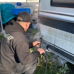 Adam Maksylewicz - Czyszczenie przyczepy kempingowej myjką ciśnieniową. Mężczyzna w czarnej bluzie z kapturem czyści bok przyczepy, z której unosi się para. Widoczne trawa i pojemniki.