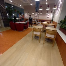 Adam Maksylewicz - Pracownik na drabinie czyści oświetlenie w kawiarni. Widoczne stoliki, krzesła i elementy wystroju wnętrza. Ujęcie z perspektywy podłogi, podkreślające wysokość pomieszczenia.