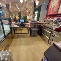 Adam Maksylewicz - Wnętrze kawiarni Costa Coffee z widocznym wyposażeniem: ekspres do kawy, lada chłodnicza, kasa fiskalna i pracownik na drabinie przy dekoracji roślinnej.