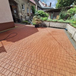 Adam Maksylewicz - Czysta, brązowa kostka brukowa po umyciu w Szczecinie. Widoczne budynki w tle i wąż do mycia. Świeżo umyta powierzchnia kostki.