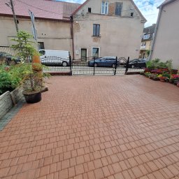 Adam Maksylewicz - Czerwona kostka brukowa na podjeździe przed domem, widok z góry. W tle brama, zaparkowane auta i budynki. Po bokach rośliny w donicach i małe drzewko.