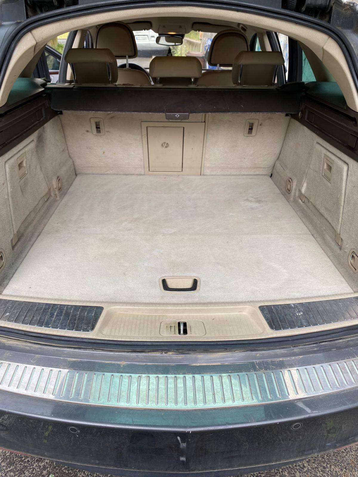 Wnętrze bagażnika kombi z beżową tapicerką, widoczne ślady użytkowania i zabrudzenia na wykładzinie. Klapa bagażnika częściowo otwarta.