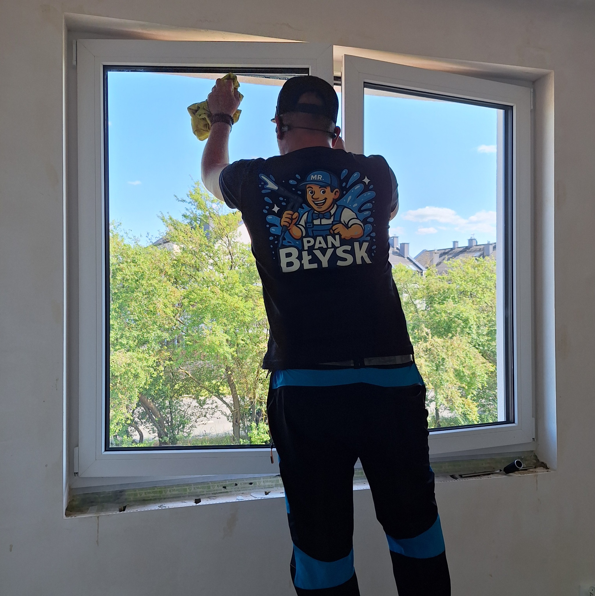 Pracownik w Szczecinie myje okno, widok z tyłu. Na koszulce logo firmy sprzątającej. Za oknem zieleń drzew i błękit nieba. Wnętrze w trakcie wykończenia.