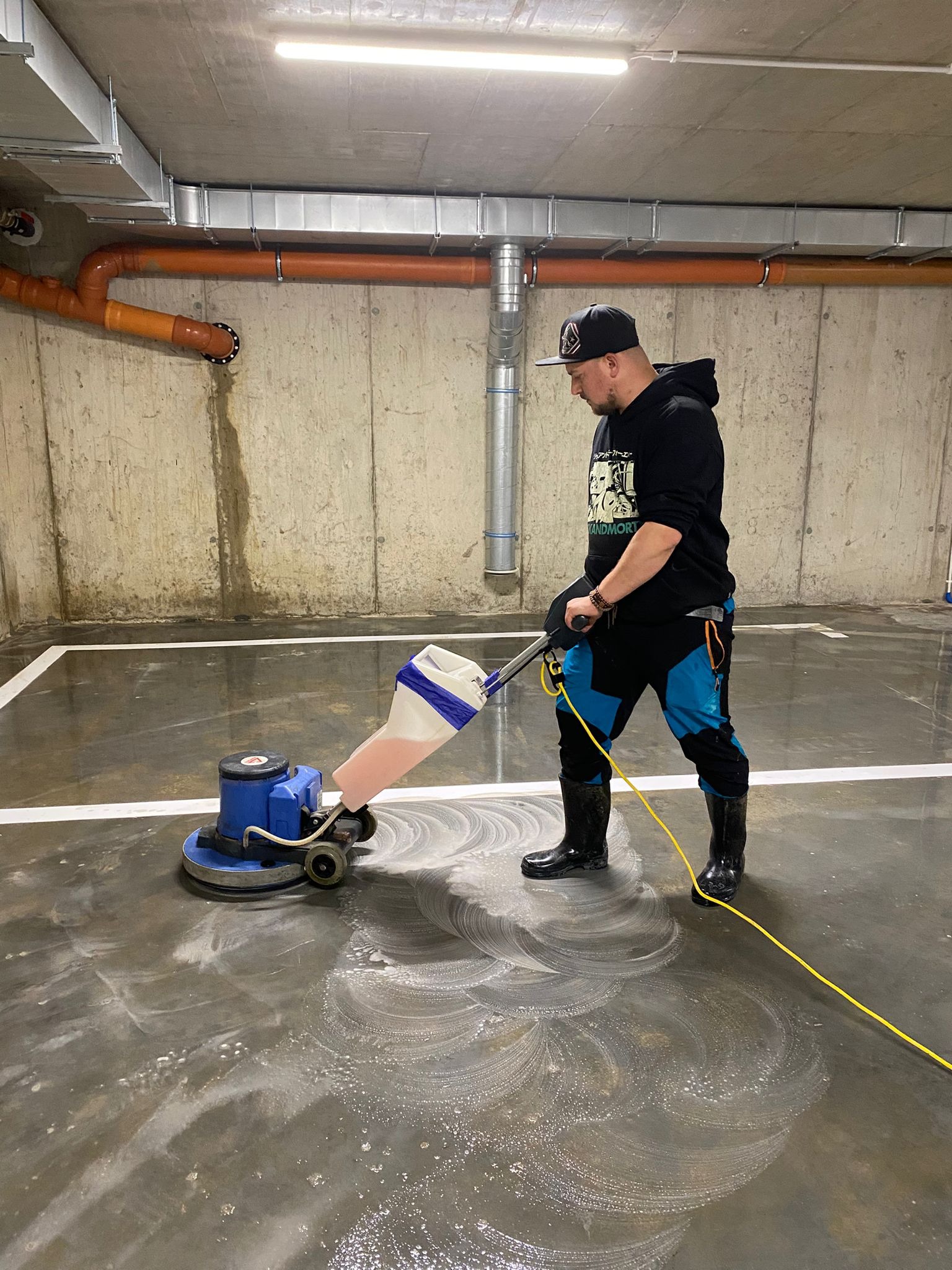 Mężczyzna w czapce i gumowcach czyści posadzkę w garażu maszyną do szorowania. Widoczne ślady piany i czyszczona powierzchnia. Pomarańczowe rury na ścianie w tle.