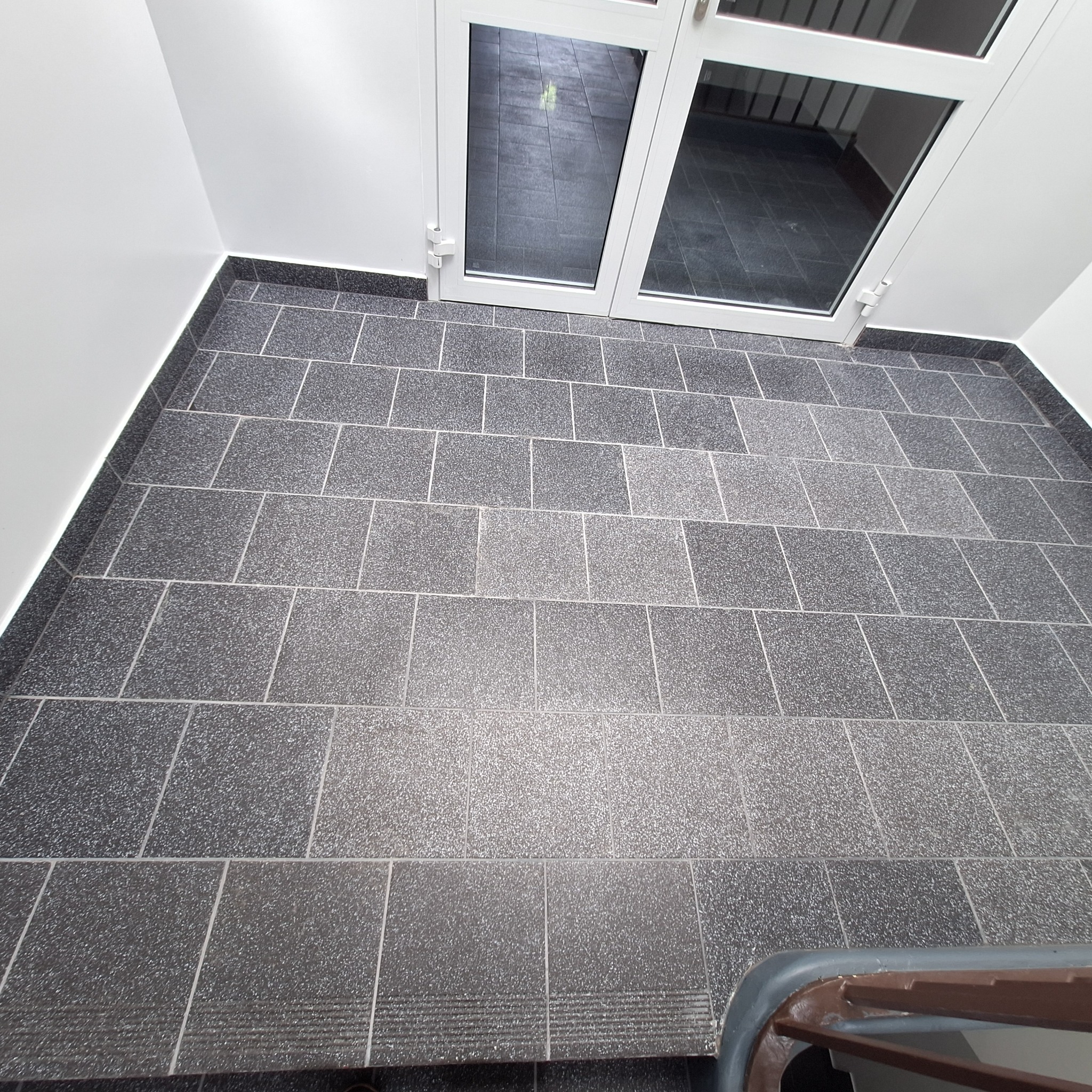 Szara kostka brukowa ułożona przed białymi drzwiami wejściowymi, widok z góry, z delikatnym fragmentem poręczy schodów w dolnym rogu kadru.