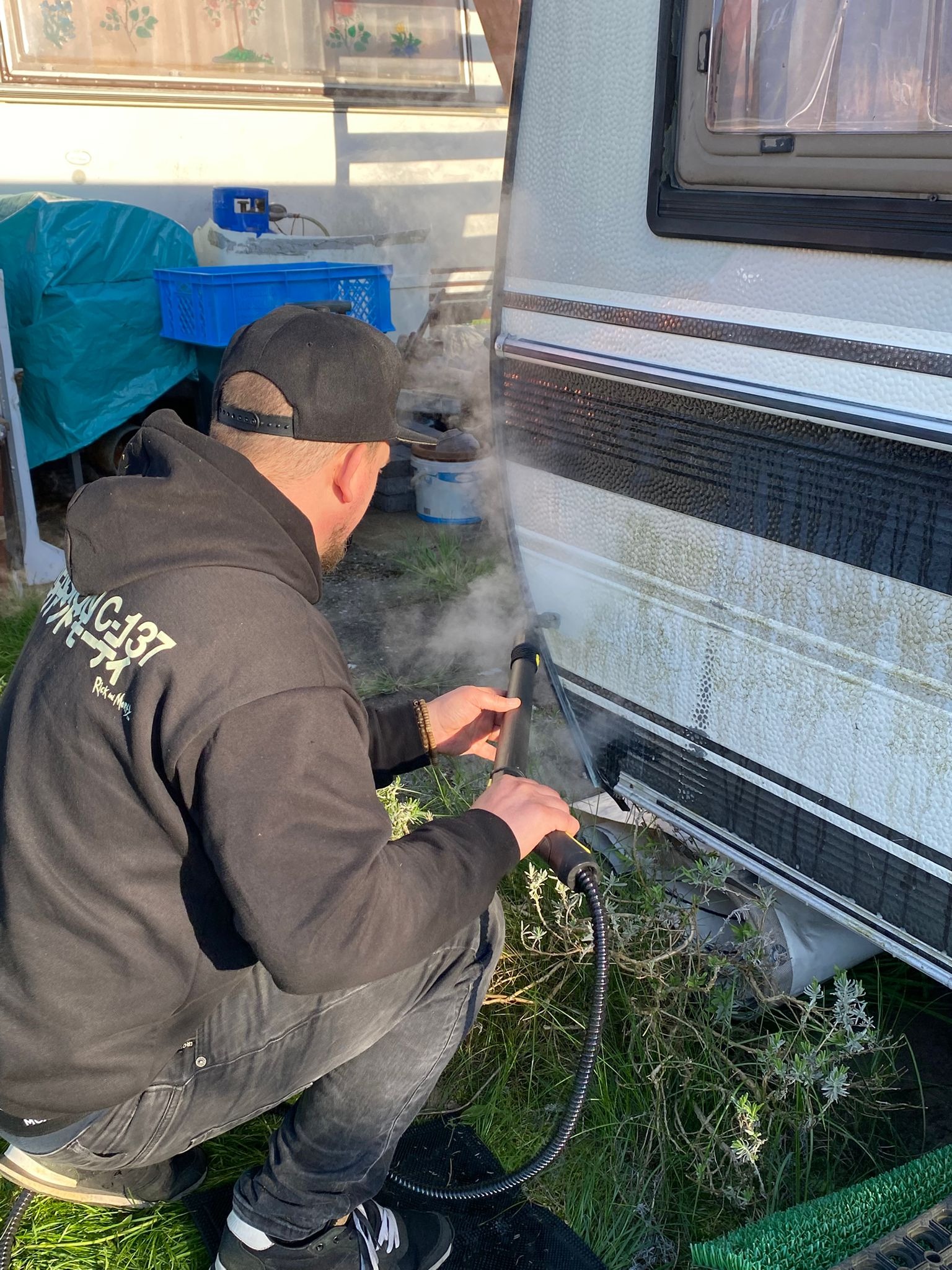 Czyszczenie przyczepy kempingowej myjką ciśnieniową. Mężczyzna w czarnej bluzie z kapturem czyści bok przyczepy, z której unosi się para. Widoczne trawa i pojemniki.