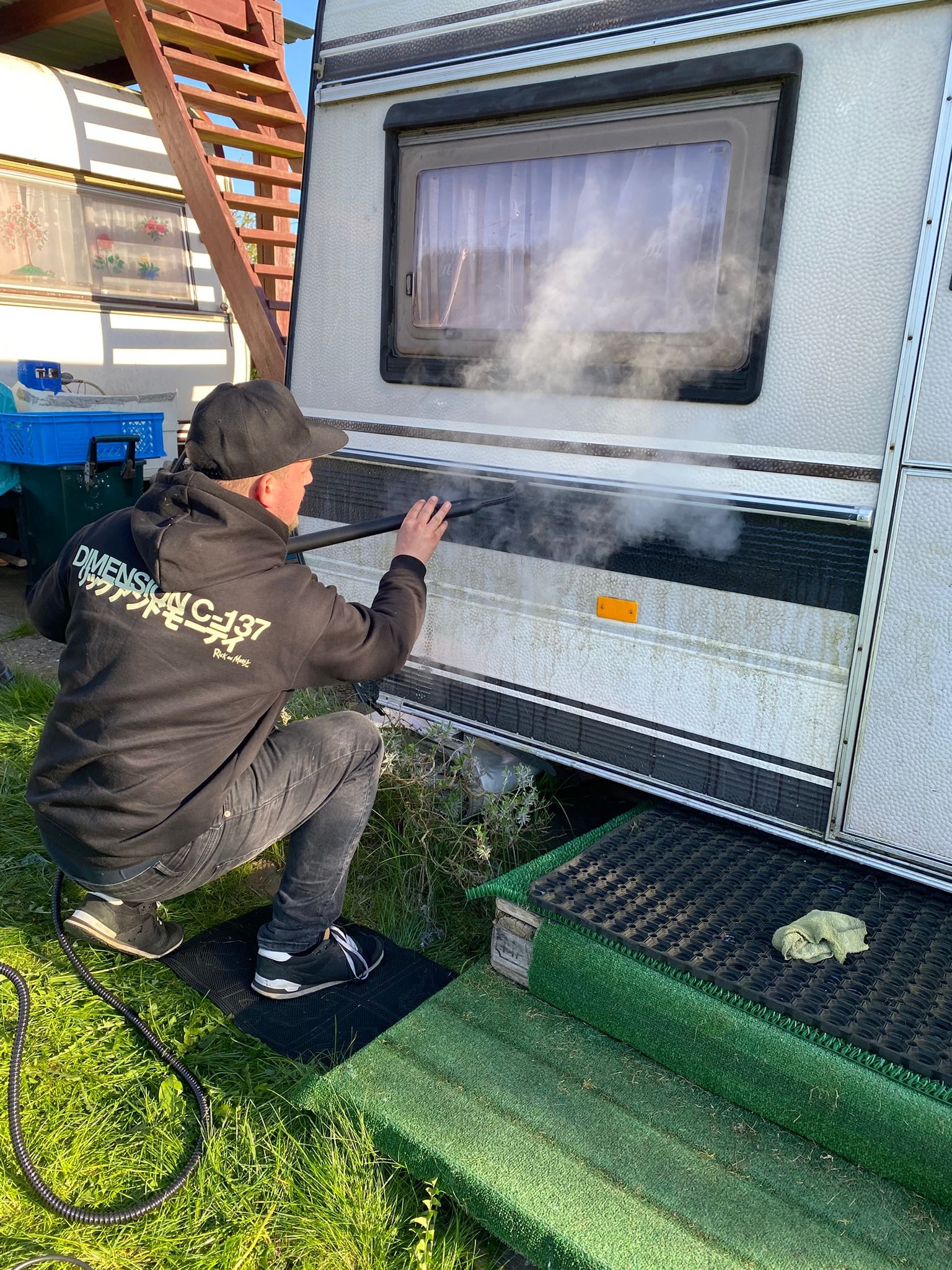 Czyszczenie parowe przyczepy kempingowej: mężczyzna w czarnej bluzie usuwa zabrudzenia z boku pojazdu, używając parownicy. Widoczna para wodna i detale przyczepy.