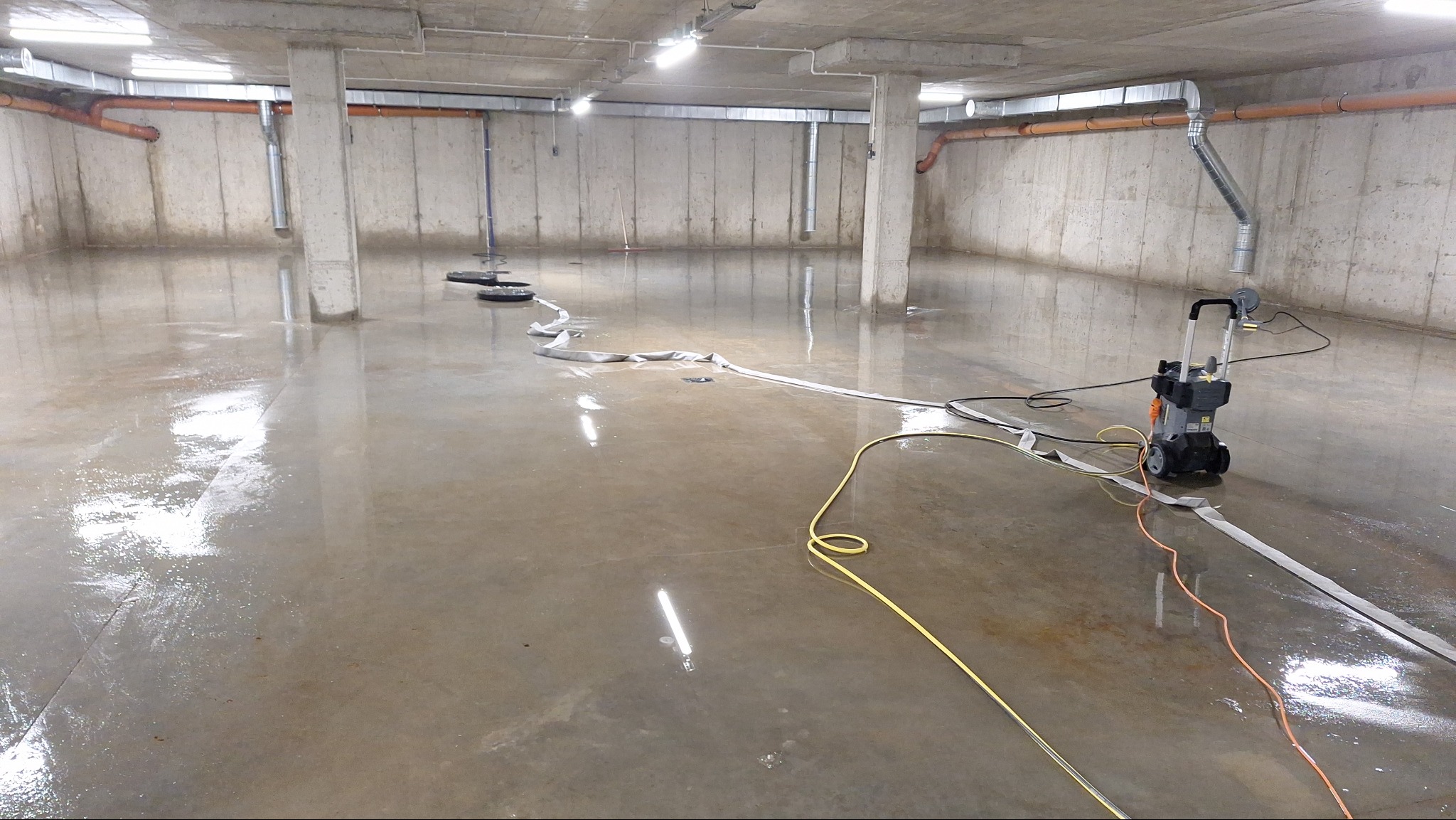 Wnętrze hali z betonową posadzką po myciu. Widoczna myjka ciśnieniowa, węże i odpływy. Świeżo umyta podłoga odbija światło lamp.