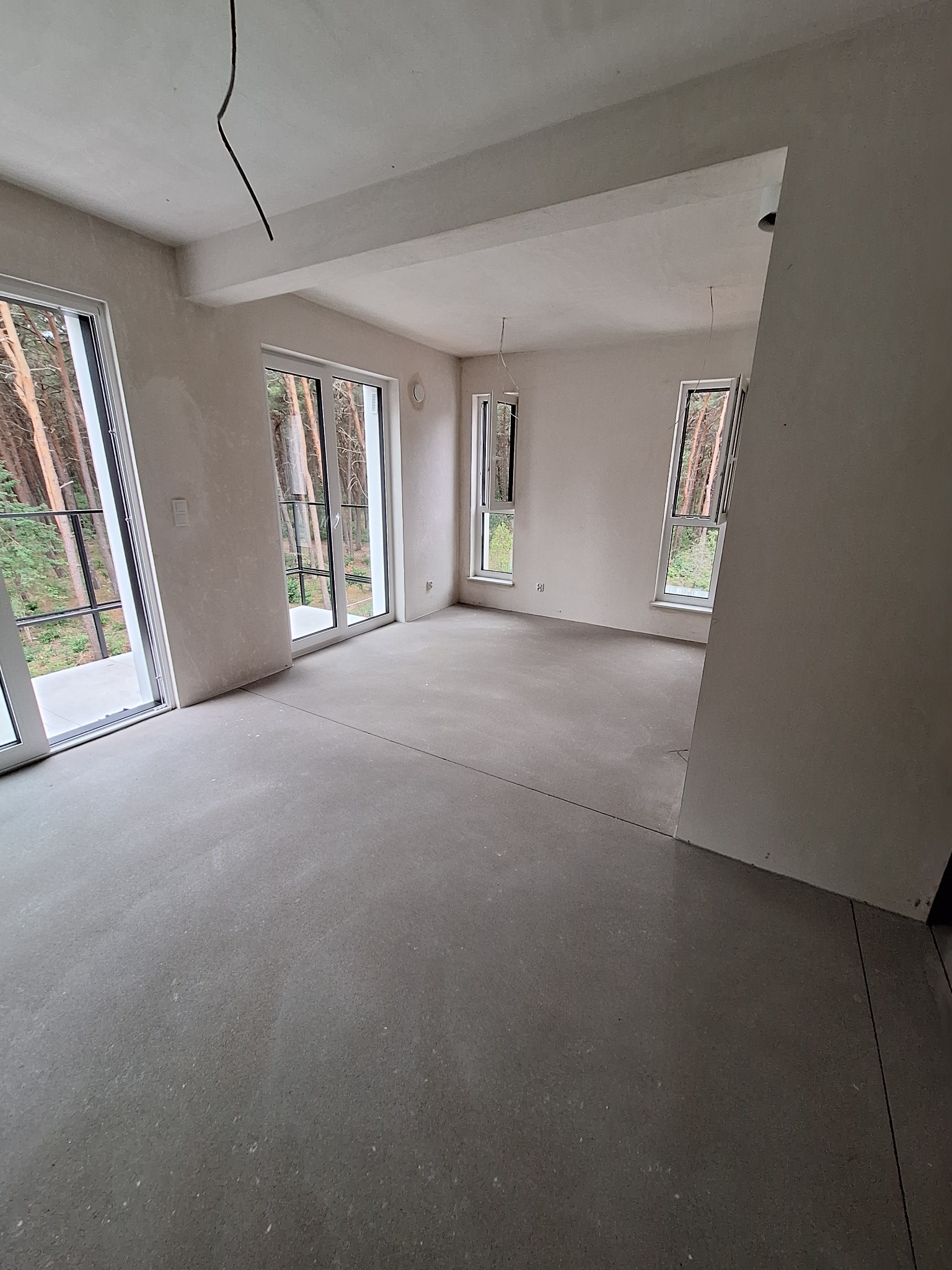 Wnętrze w stanie deweloperskim z surową posadzką, widok na las przez duże okna balkonowe. Minimalistyczna przestrzeń z widocznymi przewodami elektrycznymi.