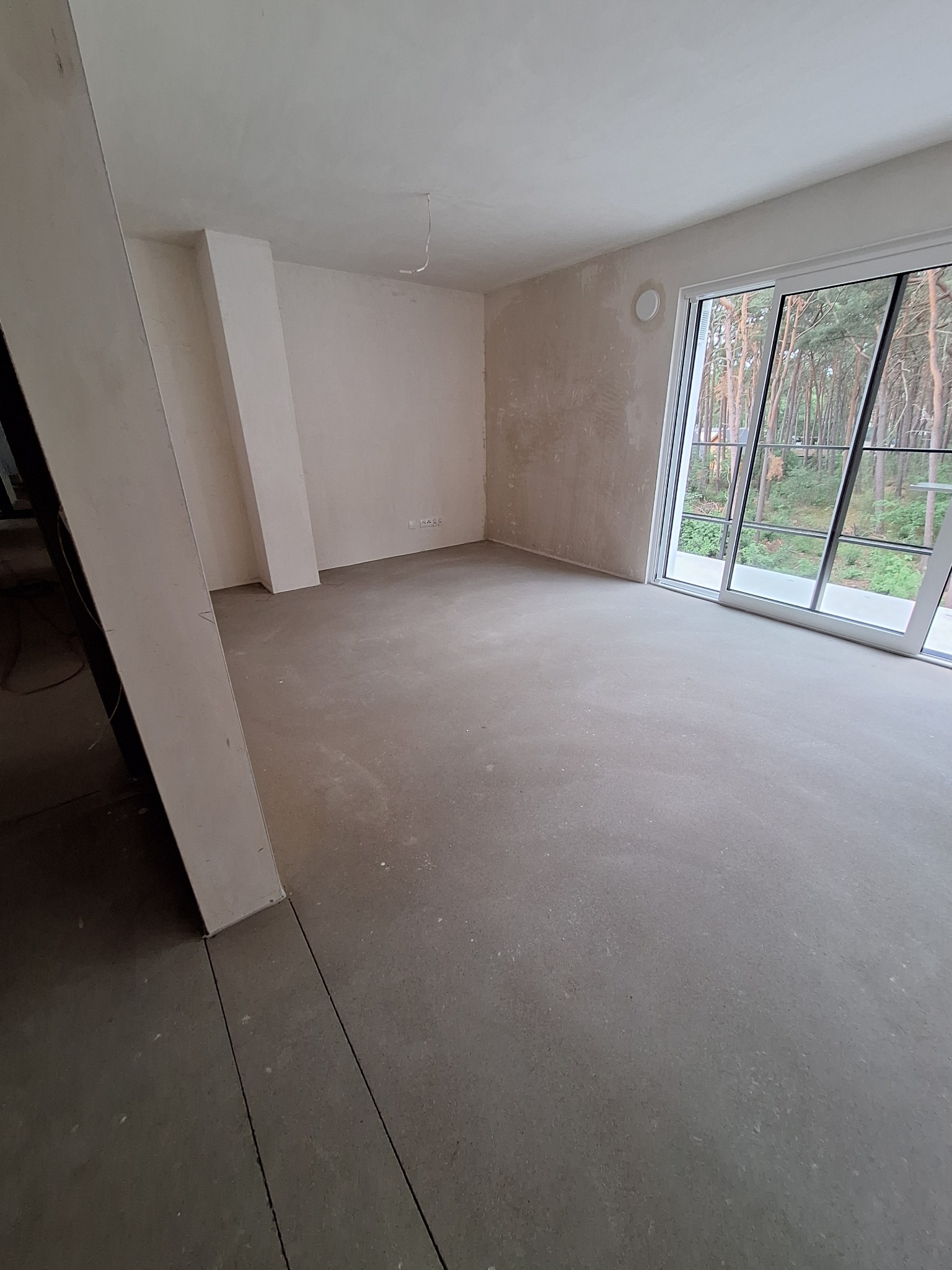 Wnętrze w stanie deweloperskim z surowymi ścianami i wylewką, duże okno przesuwne z widokiem na drzewa. Widoczne ślady po pracach wykończeniowych.