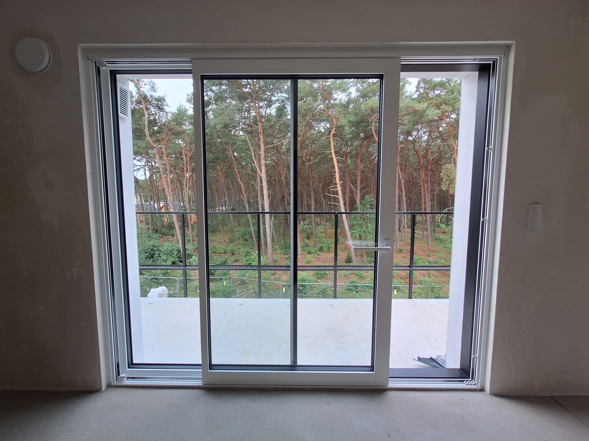 Nowe, przesuwne okno balkonowe z widokiem na las sosnowy. Białe ramy, czarne profile. Świeżo wylana posadzka, ściany w stanie surowym. Minimalistyczny design.