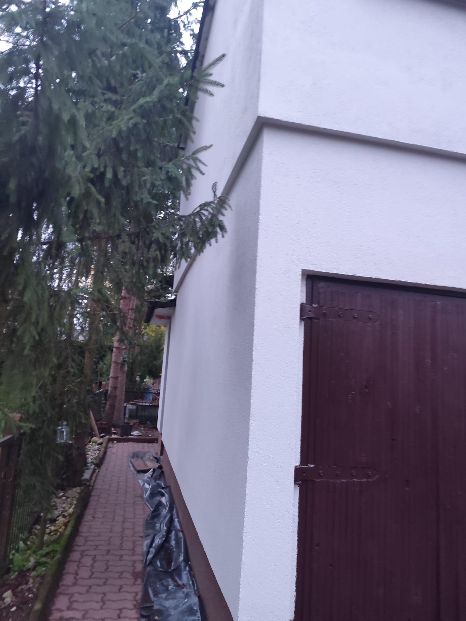 Boczny widok na świeżo pomalowaną, białą elewację budynku z brązowymi drzwiami garażowymi, z fragmentem zielonego drzewa iglastego po lewej stronie.