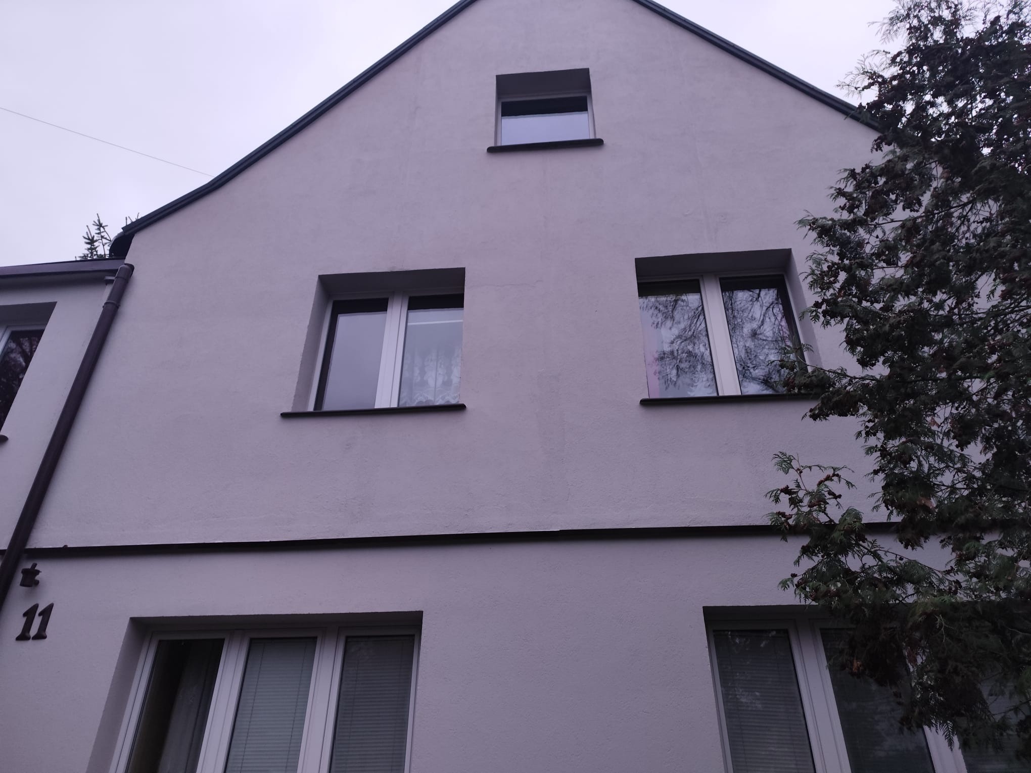 Jasna elewacja budynku z trzema rzędami okien, numer domu '11' po lewej stronie, fragment drzewa po prawej. Ujęcie z dołu, pochmurne niebo w tle.
