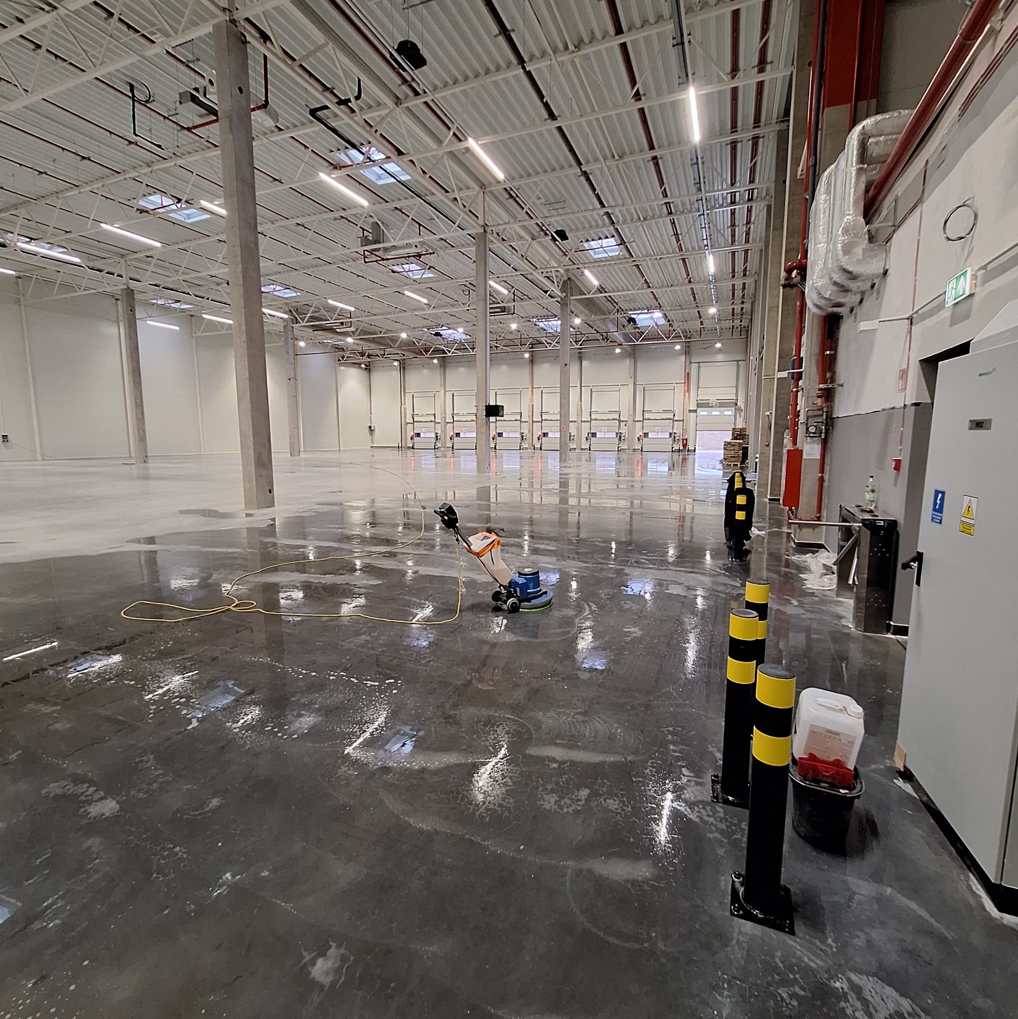 Maszyna czyszcząca posadzkę w hali magazynowej. Widoczne słupy, bramy wjazdowe i akcesoria do czyszczenia. Podłoga mokra, w trakcie sprzątania.