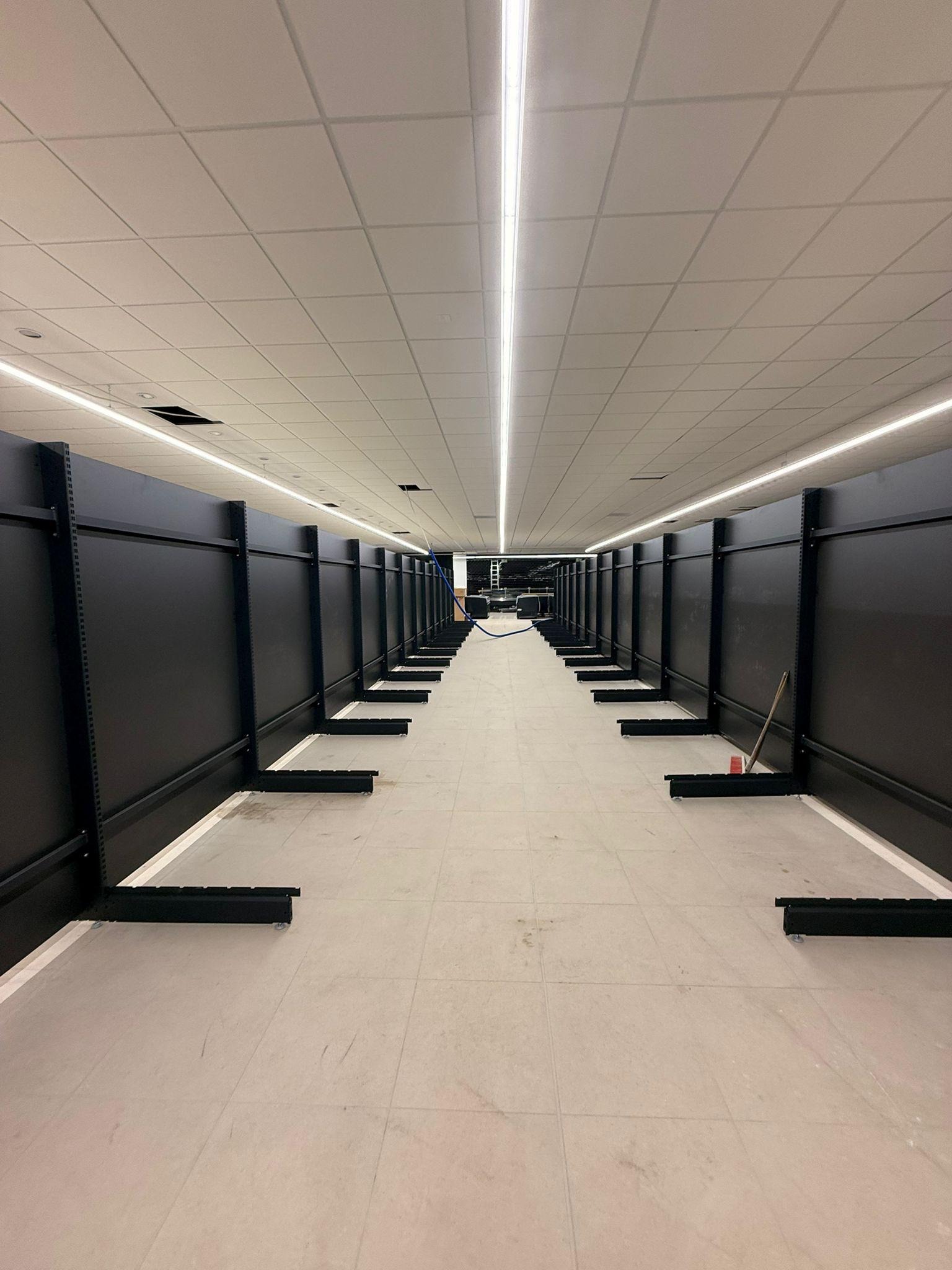 Montaż czarnych regałów w długim, jasnym pomieszczeniu z płytkami podłogowymi. Widoczna drabina w oddali. Oświetlenie liniowe sufitowe.