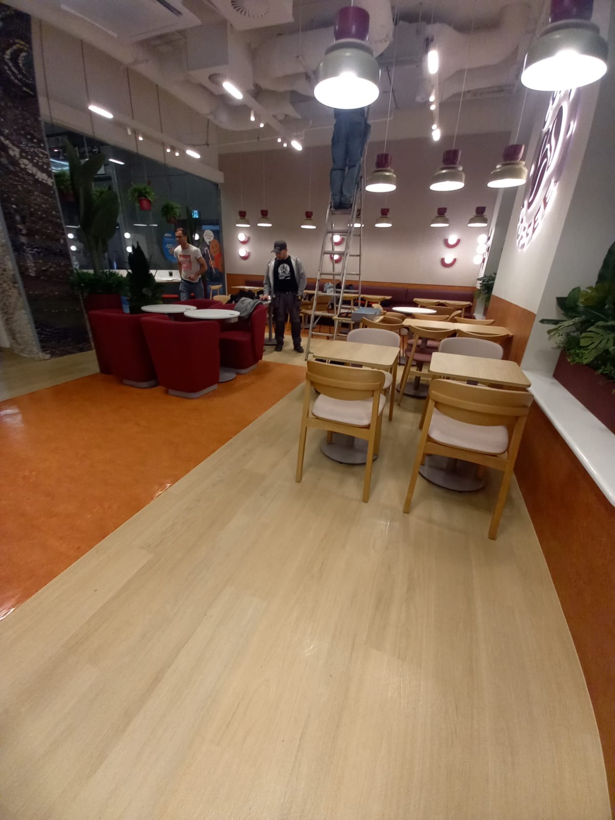 Pracownik na drabinie czyści oświetlenie w kawiarni. Widoczne stoliki, krzesła i elementy wystroju wnętrza. Ujęcie z perspektywy podłogi, podkreślające wysokość pomieszczenia.