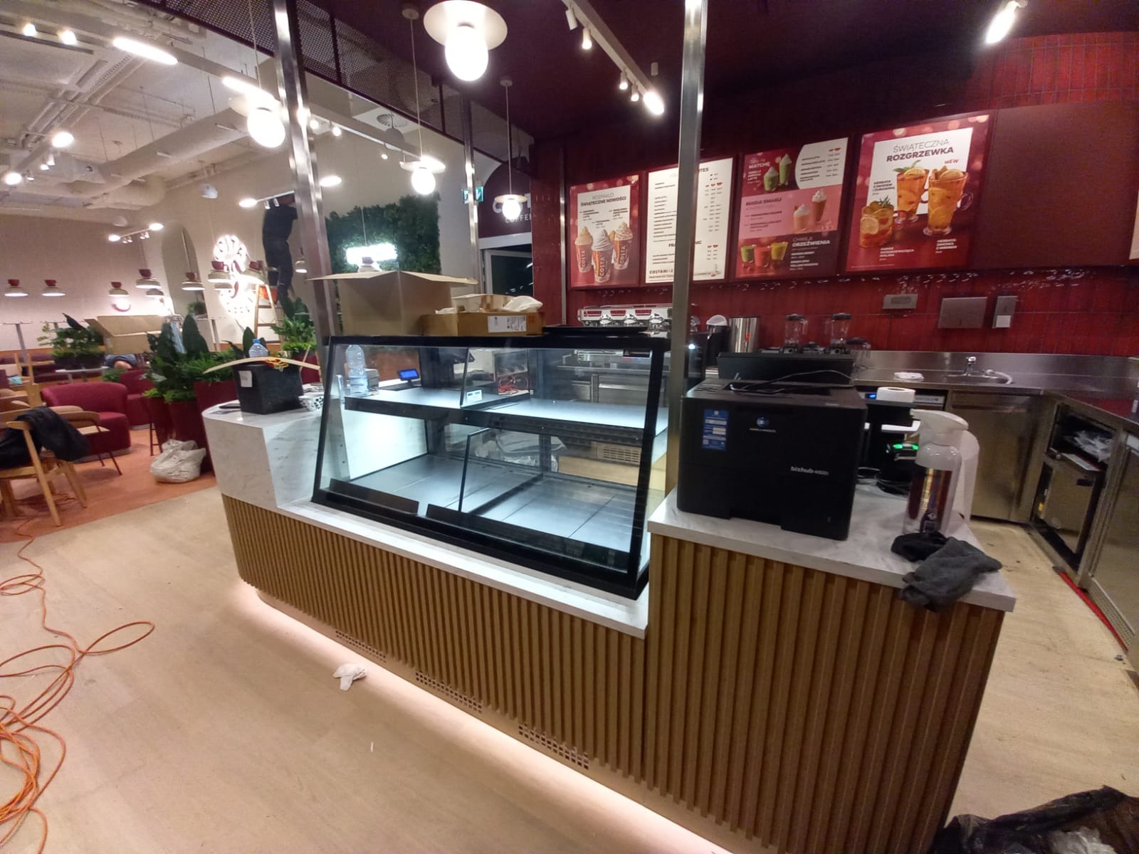 Pusta witryna chłodnicza w kawiarni, z widocznymi plakatami menu na ścianie i elementami wyposażenia. Widok na wnętrze lokalu z miejscami siedzącymi.