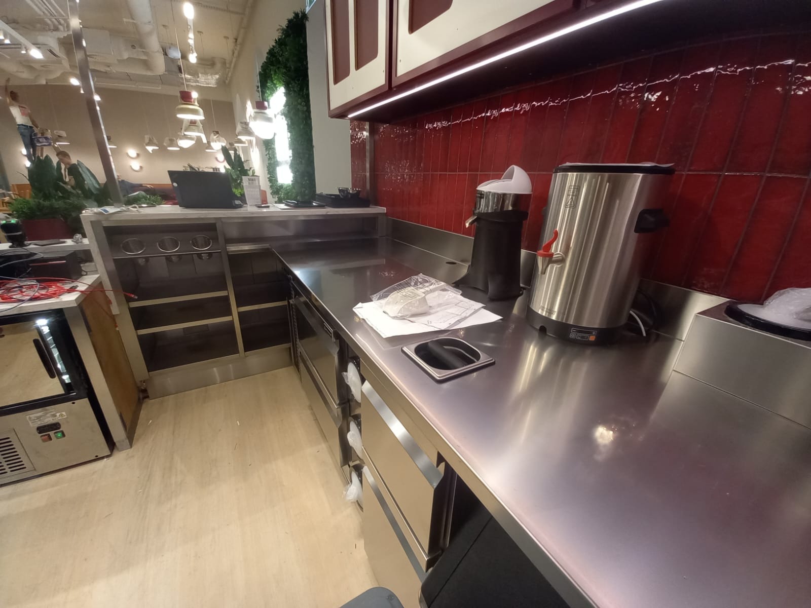Błyszcząca, metalowa powierzchnia robocza w lokalu gastronomicznym z dozownikiem do napojów i akcesoriami. Czerwone płytki na ścianie w tle. Widoczne szafki i wyposażenie.
