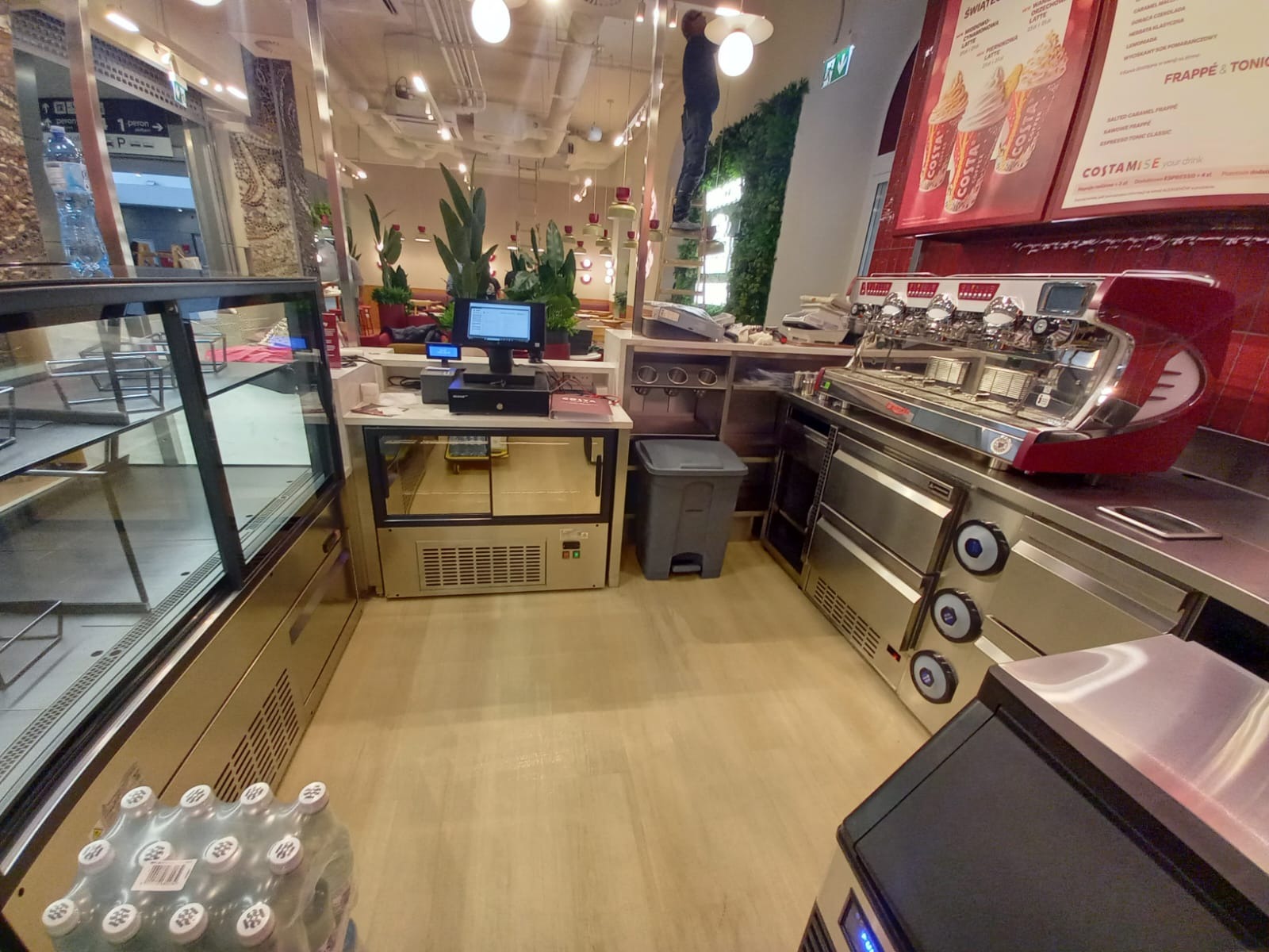 Wnętrze kawiarni Costa Coffee z widocznym wyposażeniem: ekspres do kawy, lada chłodnicza, kasa fiskalna i pracownik na drabinie przy dekoracji roślinnej.