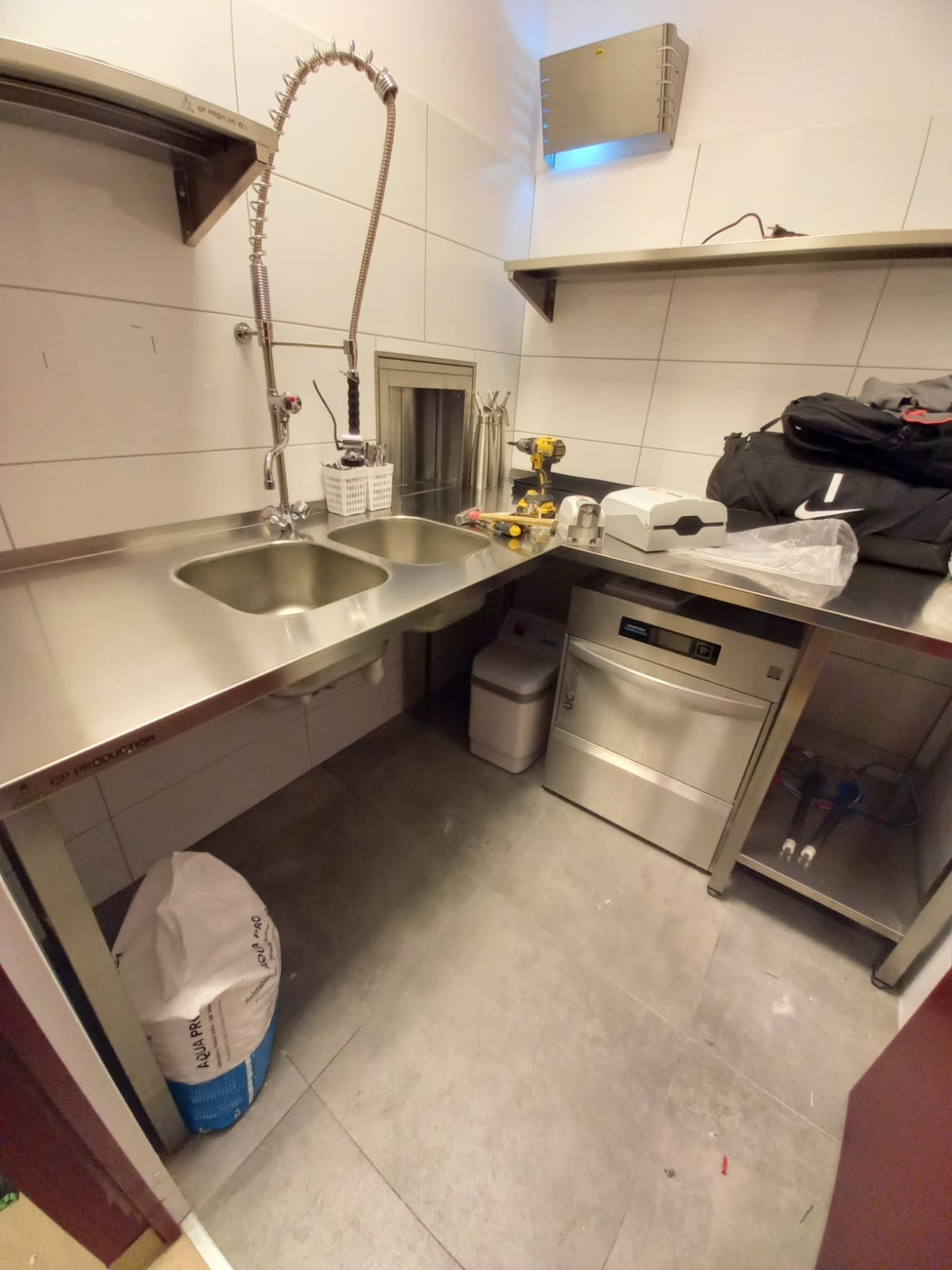 Profesjonalna kuchnia ze stali nierdzewnej: dwukomorowy zlew z elastyczną wylewką, zmywarka gastronomiczna, narzędzia i akcesoria. Widok na czyste, kafelkowane ściany.