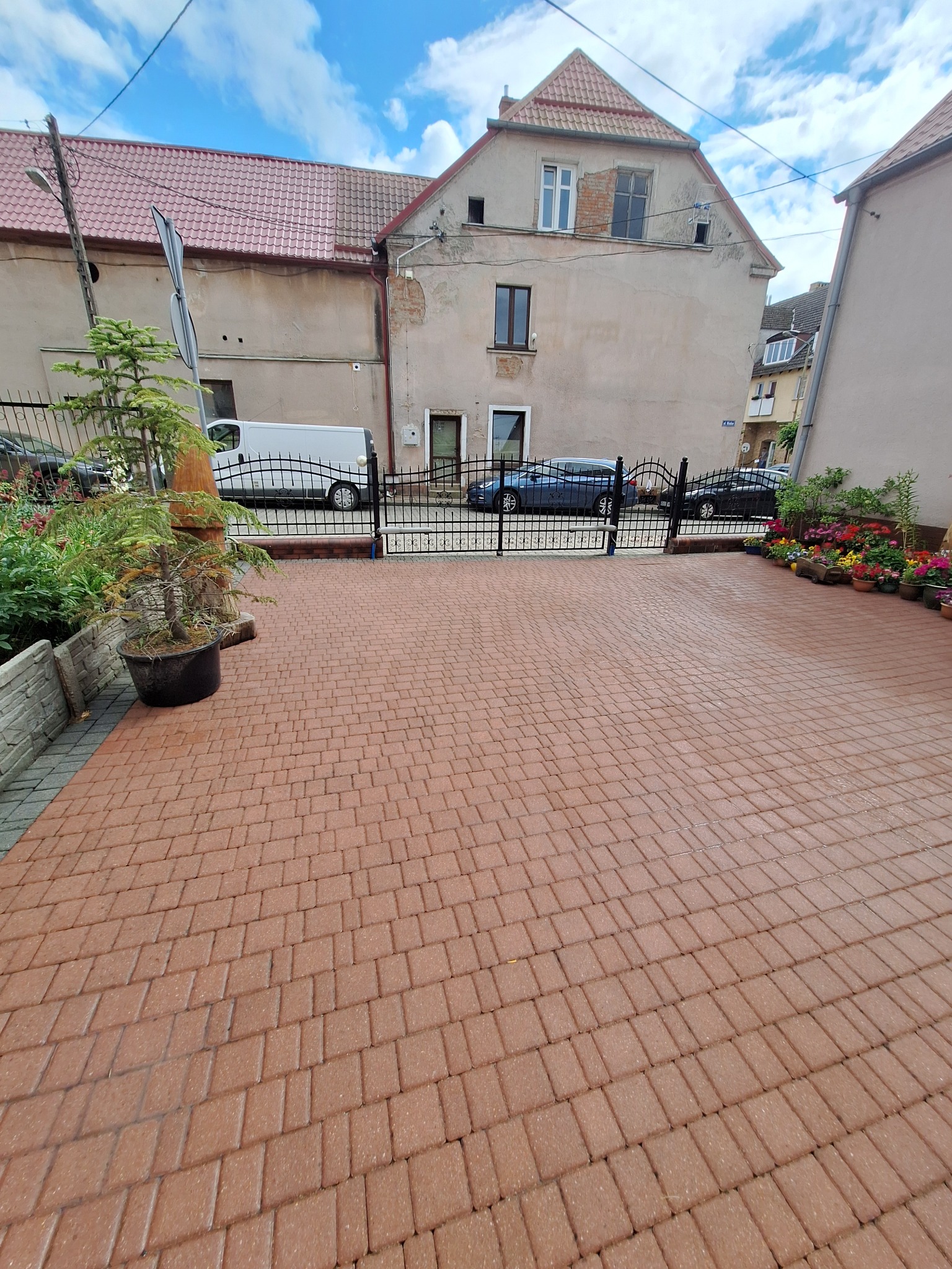 Czerwona kostka brukowa na podjeździe przed domem, widok z góry. W tle brama, zaparkowane auta i budynki. Po bokach rośliny w donicach i małe drzewko.