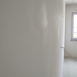 Profesjonalne wykończenia wnętrz Łukasz Szulc - Świeżo położona, jasna, prawie gładka ściana we Wrocławiu, z widocznymi przewodami elektrycznymi wystającymi ze ściany przy podłodze, w tle okno i surowy stan wykończenia.