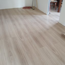 Profesjonalne wykończenia wnętrz Łukasz Szulc - Nowo położona podłoga laminowana o jasnym, drewnopodobnym wzorze w pustym pokoju z białymi ścianami; widoczne narzędzia i zwinięty przedłużacz w rogu, sugerujące trwające prace wykończeniowe.