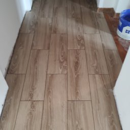 Profesjonalne wykończenia wnętrz Łukasz Szulc - Ułożona podłoga z płytek imitujących drewno w przejściu między pomieszczeniami, widoczna listwa progowa i fragment wiadra z farbą.