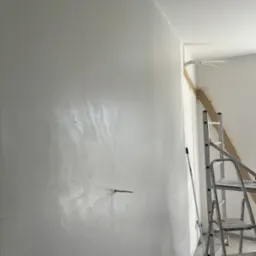 Ściana po nałożeniu gładzi szpachlowej, widoczne delikatne refleksy światła, w rogu drabina malarska i wystające kable elektryczne.