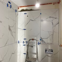 Profesjonalne wykończenia wnętrz Łukasz Szulc - Ściana łazienki w trakcie układania białych płytek imitujących marmur, z użyciem niebieskich klipsów poziomujących, widoczna drabina malarska i wystająca żarówka.