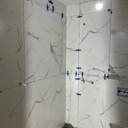 Profesjonalne wykończenia wnętrz Łukasz Szulc - Świeżo ułożone, białe płytki imitujące marmur na ścianach łazienki, z użyciem niebieskich klipsów poziomujących, widoczny fragment instalacji sanitarnej i surowy stan wykończenia.