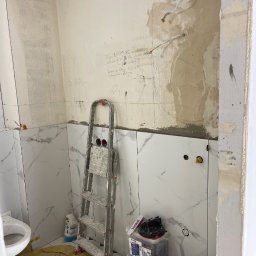 Profesjonalne wykończenia wnętrz Łukasz Szulc - Łazienka w trakcie remontu: częściowo ułożone białe płytki imitujące marmur na ścianach, widoczne ślady po instalacjach elektrycznych, drabina malarska, pojemniki z zaprawą, przygotowane podłoże...